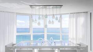 Un espace de repas moderne avec une table en verre et des chaises blanches, de grandes fenêtres offrant une vue sur l’océan et des luminaires suspendus.