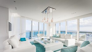 Un salon moderne avec des meubles blancs, une table basse en verre et de grandes fenêtres donnant sur l’océan.
