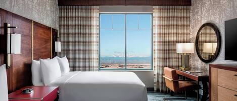 A hotel room with a large bed, a desk, a chair, a TV, and a view of mountains through the window.