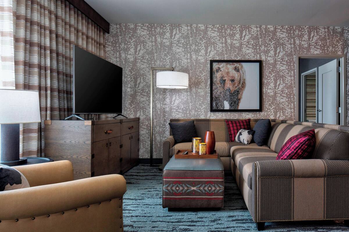 A living room with a sofa, ottoman, and television, featuring patterned wallpaper and a framed artwork.