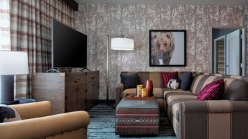A living room with a sofa, ottoman, and television, featuring patterned wallpaper and a framed artwork.