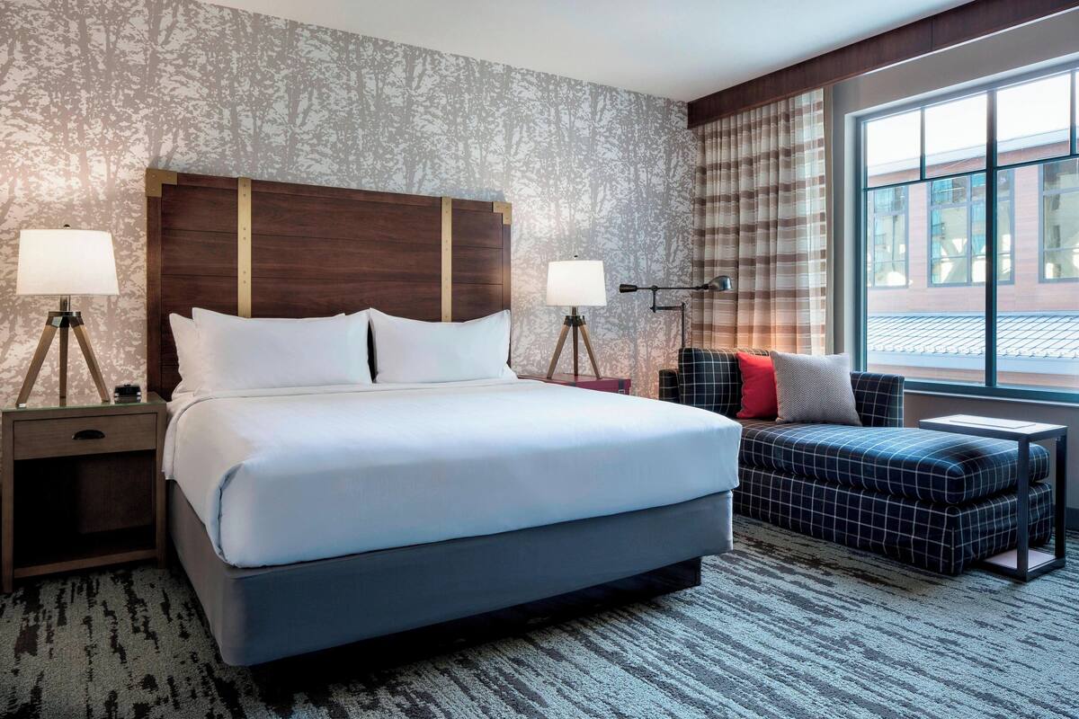 A hotel room with a large bed, two bedside tables with lamps, a sofa, and a large window with curtains.