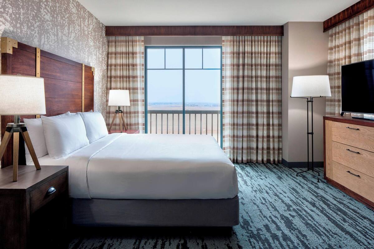 A hotel room with a large bed, a television, a desk with a lamp, and a view of the outdoors through a window.