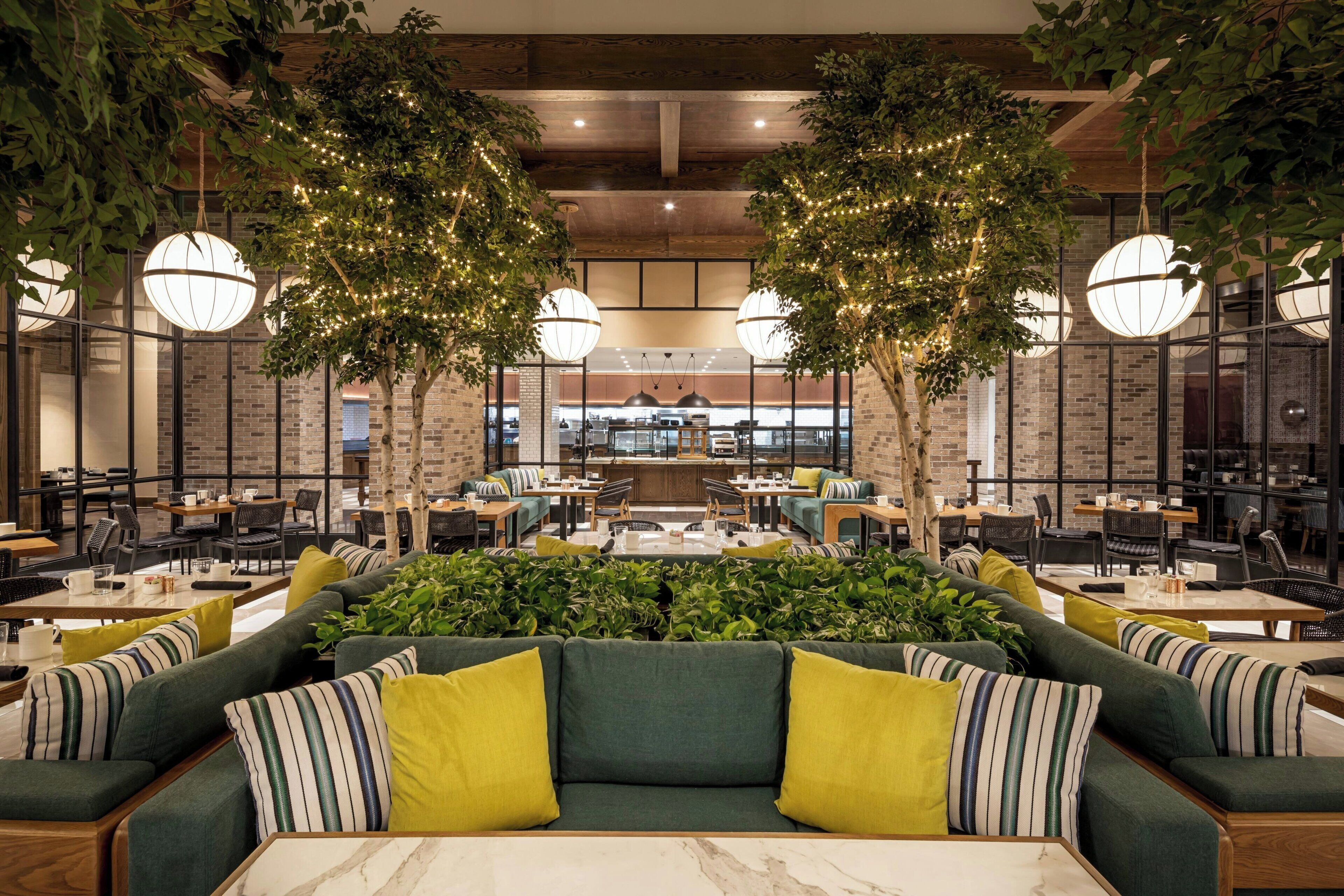 A spacious indoor area with multiple seating arrangements, including sofas and tables, surrounded by greenery and trees.