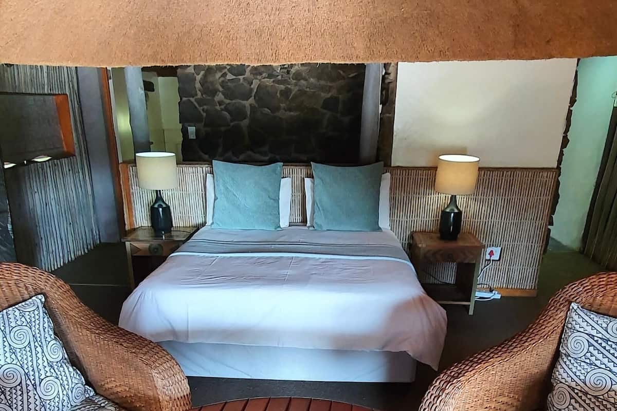 Habitación de hotel con cabecera de piedra, cama con ropa blanca, dos lámparas de noche y muebles de mimbre.