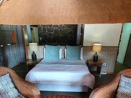 Habitación de hotel con cabecera de piedra, cama con ropa blanca, dos lámparas de noche y muebles de mimbre.