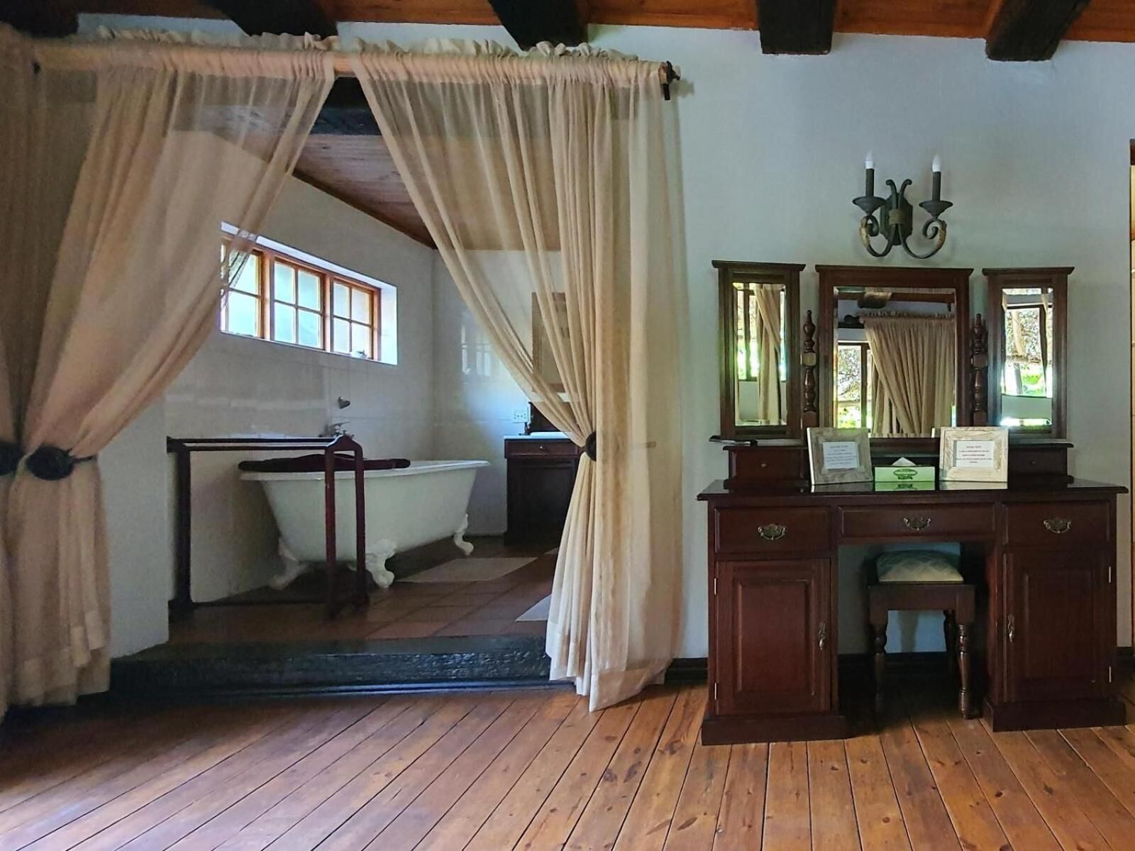A room with a wooden floor, a white bathtub, a wooden dresser with a mirror, and a window with curtains.
