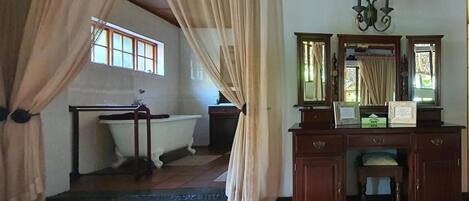 A room with a wooden floor, a white bathtub, a wooden dresser with a mirror, and a window with curtains.