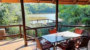 A covered outdoor seating area with a table and chairs, overlooking a lake and forested area. - Glen Afric Country Lodge (Broederstroom)