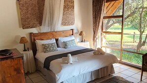A bedroom with a wooden headboard, white bedding, two bedside lamps, a nightstand, a wicker basket, a grey rug, and a large window with a view of greenery. - Glen Afric Country Lodge (Broederstroom)
