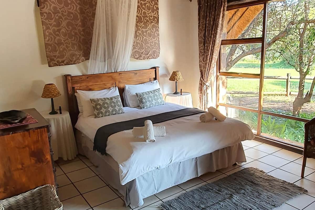 Un dormitorio con cabecera de madera, ropa de cama blanca, dos lámparas de noche, una mesita de noche, una cesta de mimbre, un tapete gris y un ventanal con vistas a la vegetación.