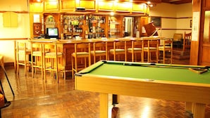Une table de billard se trouve dans une pièce dont le sol est en bois, et un coin bar est aménagé avec des tabourets et un écran d’ordinateur.