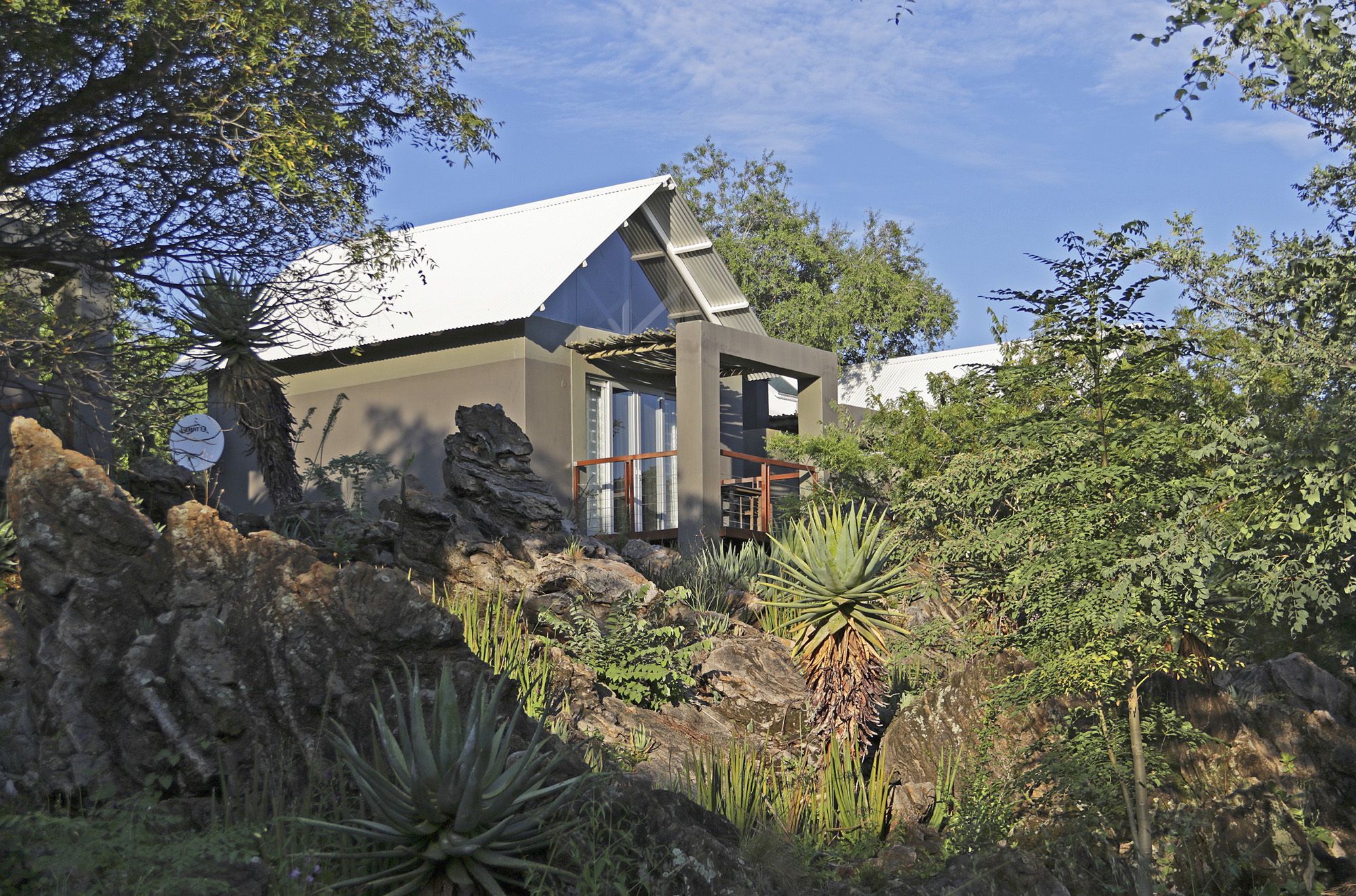 A modern house with a white roof, surrounded by natural vegetation and rocks.