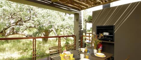 An outdoor dining area with a wooden table set for two, a grill, and a view of trees.
