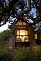 Una casa moderna de dos pisos con fachada de vidrio, rodeada de árboles y césped.