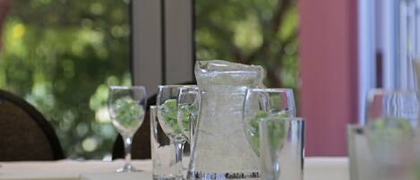 A table set with clear glasses and a pitcher, likely for water or a beverage, with a blurred green background suggesting an outdoor setting.