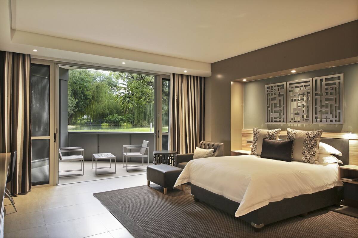 A modern bedroom with a large bed, a balcony with seating, and a view of greenery.
