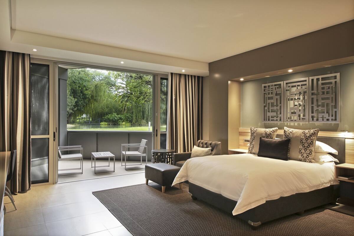 A modern bedroom with a large bed, a balcony with chairs, and a view of greenery.