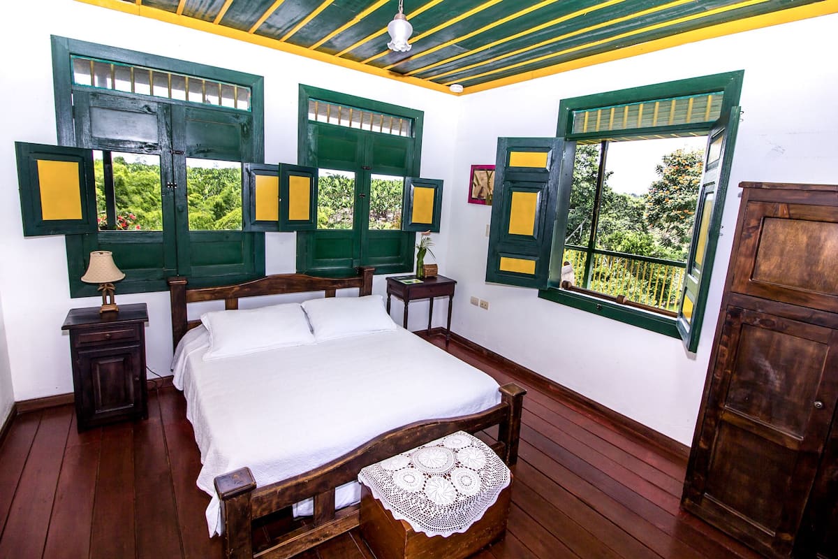 A bedroom with a wooden bed, two nightstands, a wooden wardrobe, and green-framed windows with yellow panels.