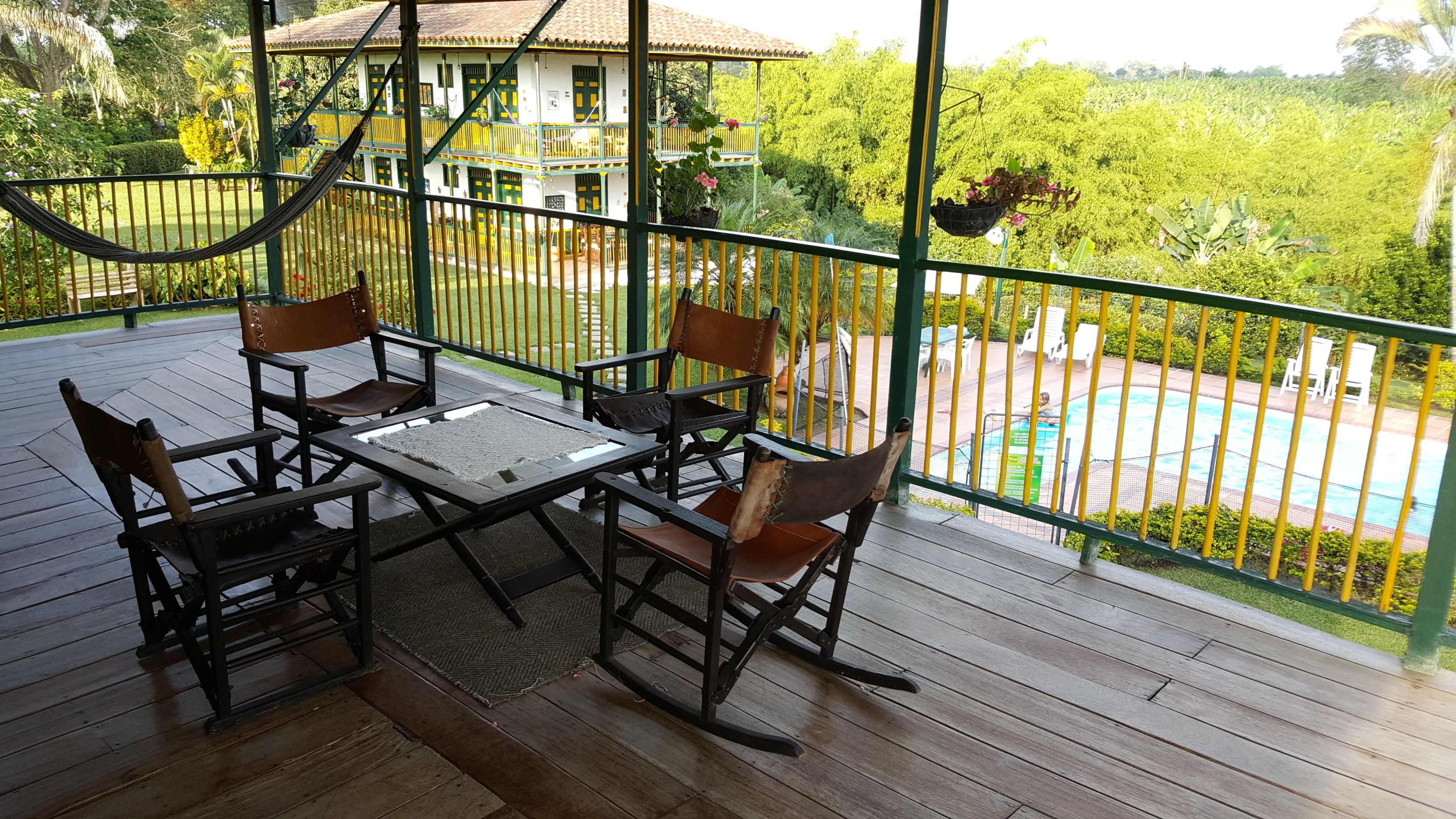 Terraza de madera con mesa y sillas, con vista a una piscina y abundante vegetación.