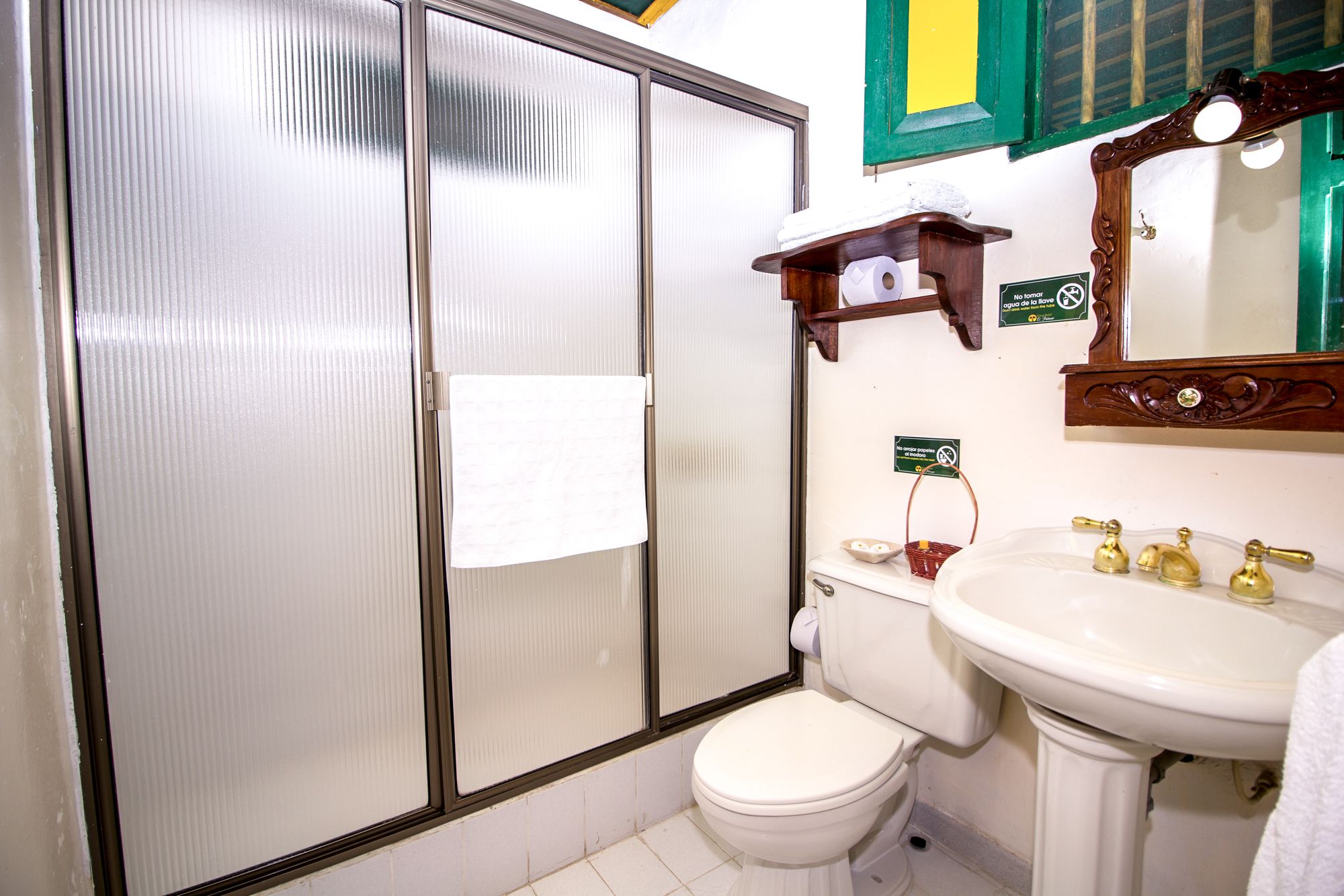 Un baño con inodoro, lavamanos y ducha. Hay carteles en la pared, una toalla colgando de la puerta de la ducha y un estante de madera sobre el inodoro.