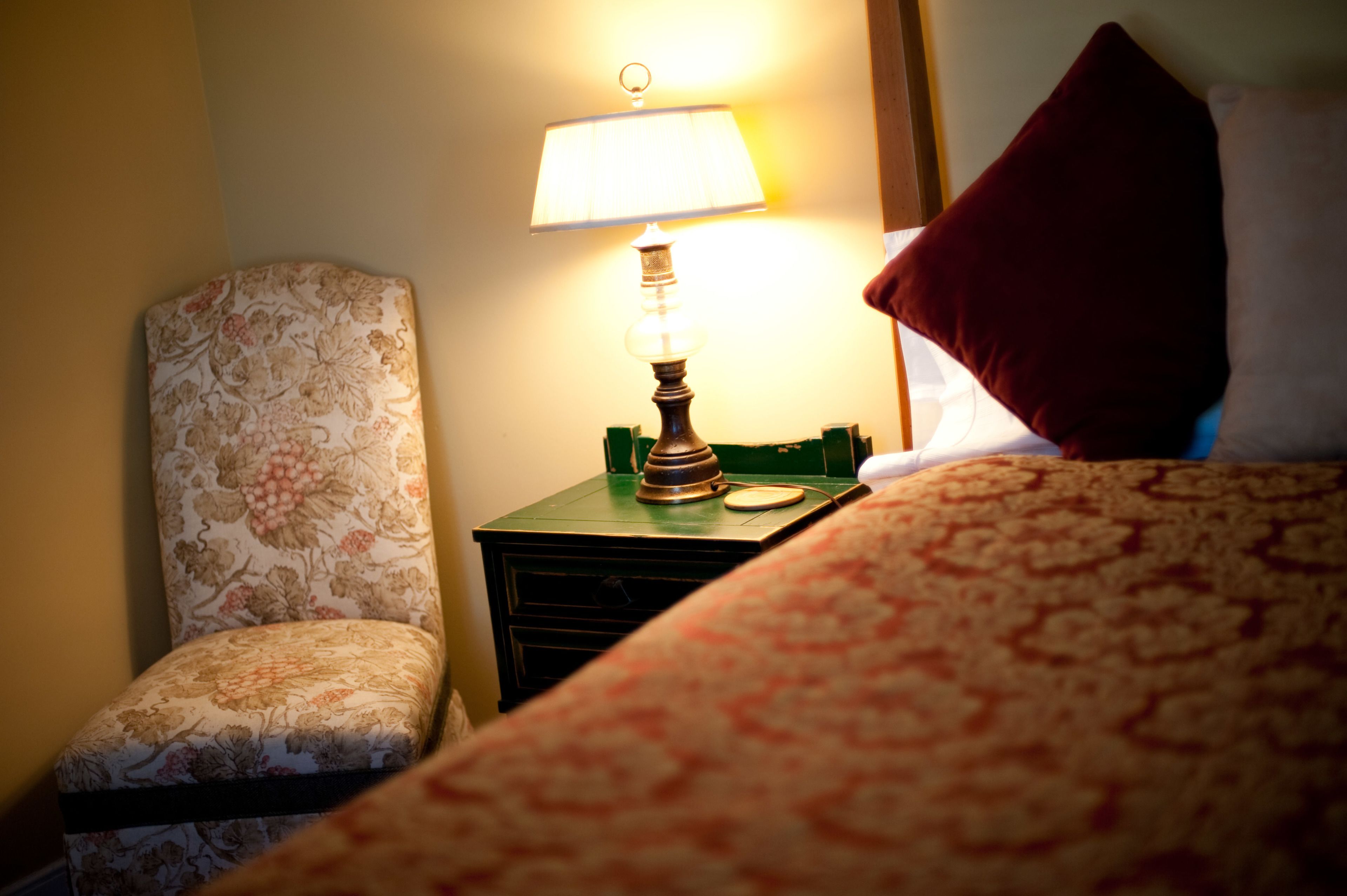 Una habitación de hotel con un sillón estampado de flores, una mesita de noche con una lámpara y una cama con colcha estampada.
