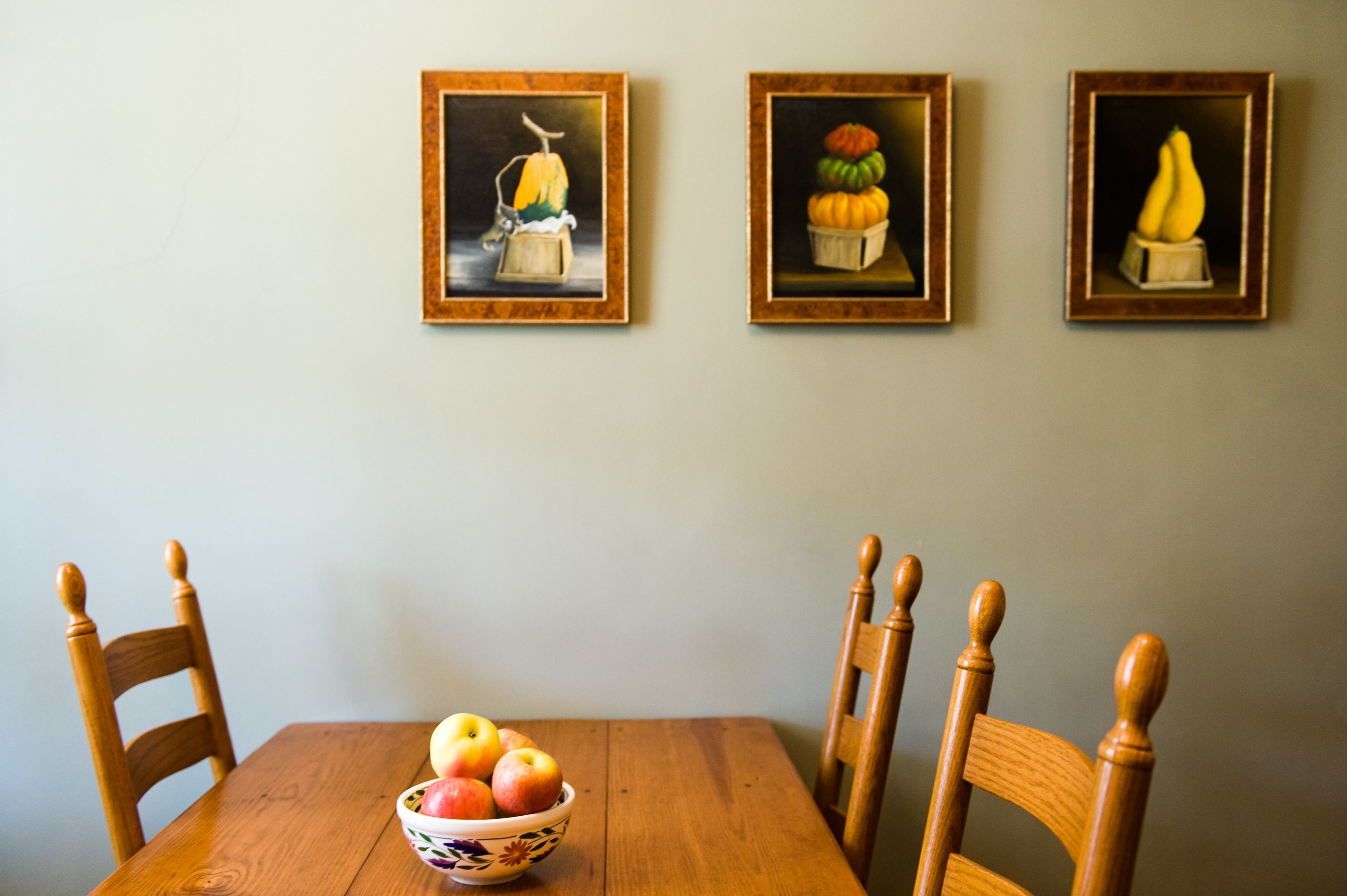 A dining table with a bowl of fruit and three framed paintings on the wall.