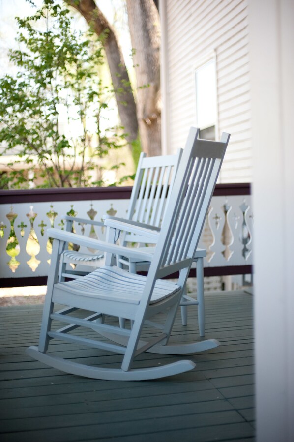 A rocking chair on a wooden deck with a white railing and a tree in the background.