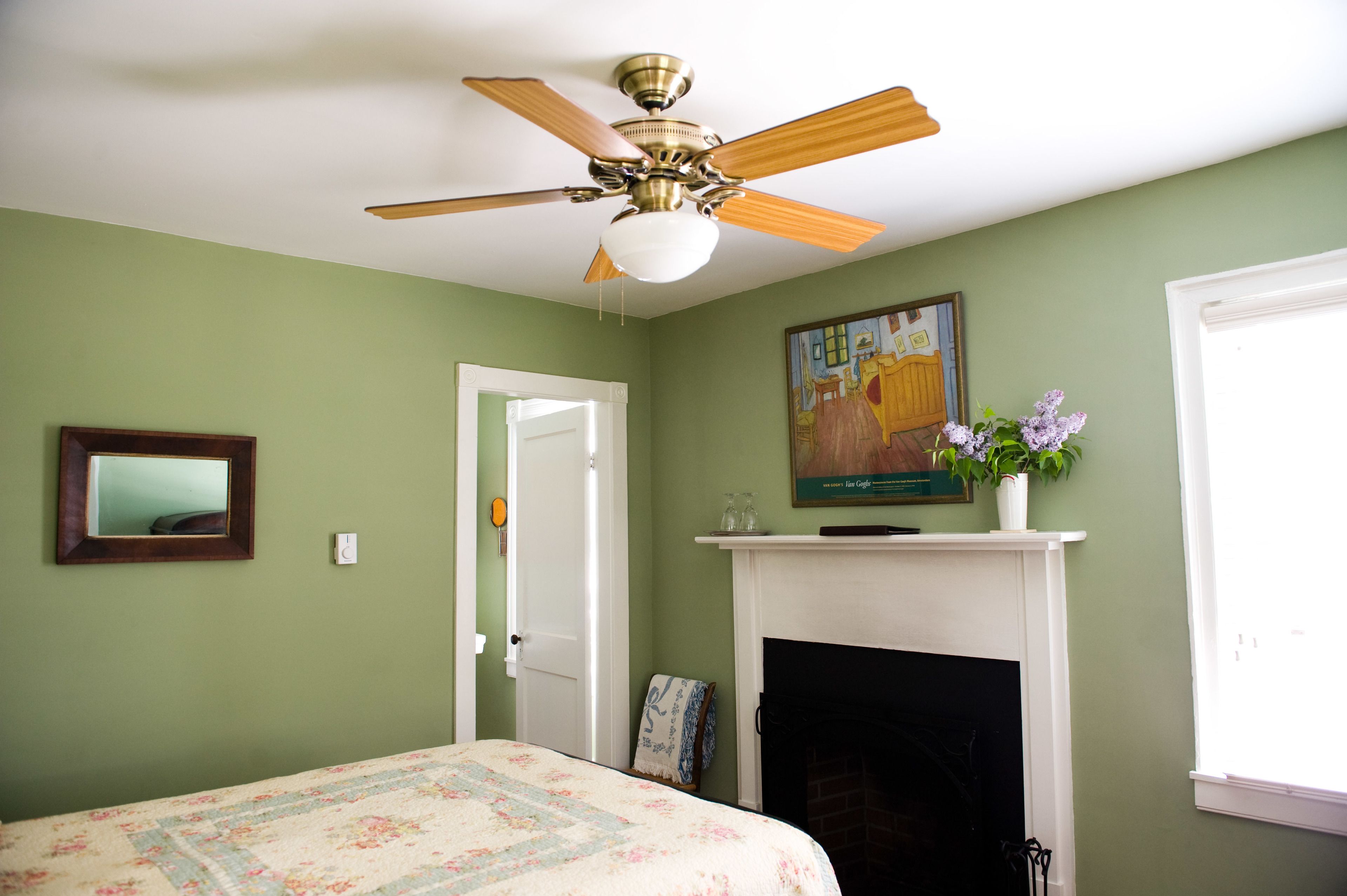 A bedroom with a bed, a ceiling fan, a fireplace, a painting, and a window.