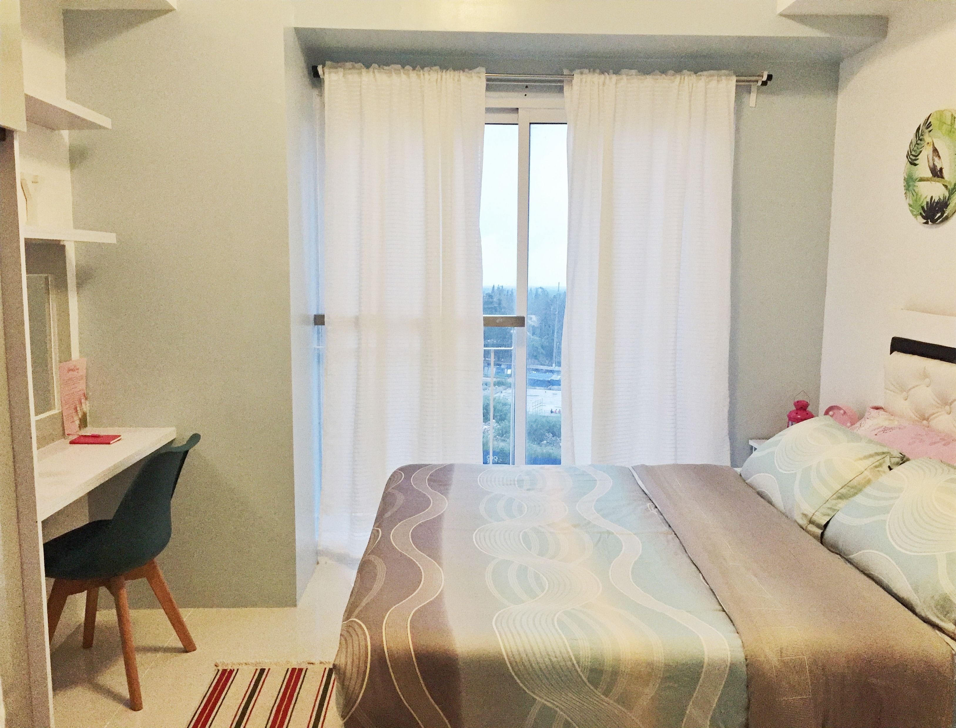 Un dormitorio con cama, escritorio y silla. Hay una ventana con cortinas y vistas al exterior.