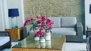 A modern living room with a glass coffee table, a grey sofa, and wicker chairs. There are decorative jars with pink flowers on the table.