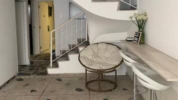 Una escalera con baranda blanca, una silla redonda con cojín y un taburete en un pasillo con piso estampado.