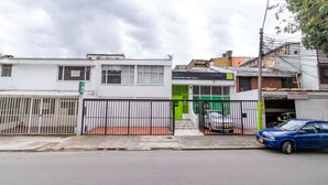 Vista de una calle con un edificio blanco, un portón verde y un coche azul estacionado a la derecha.