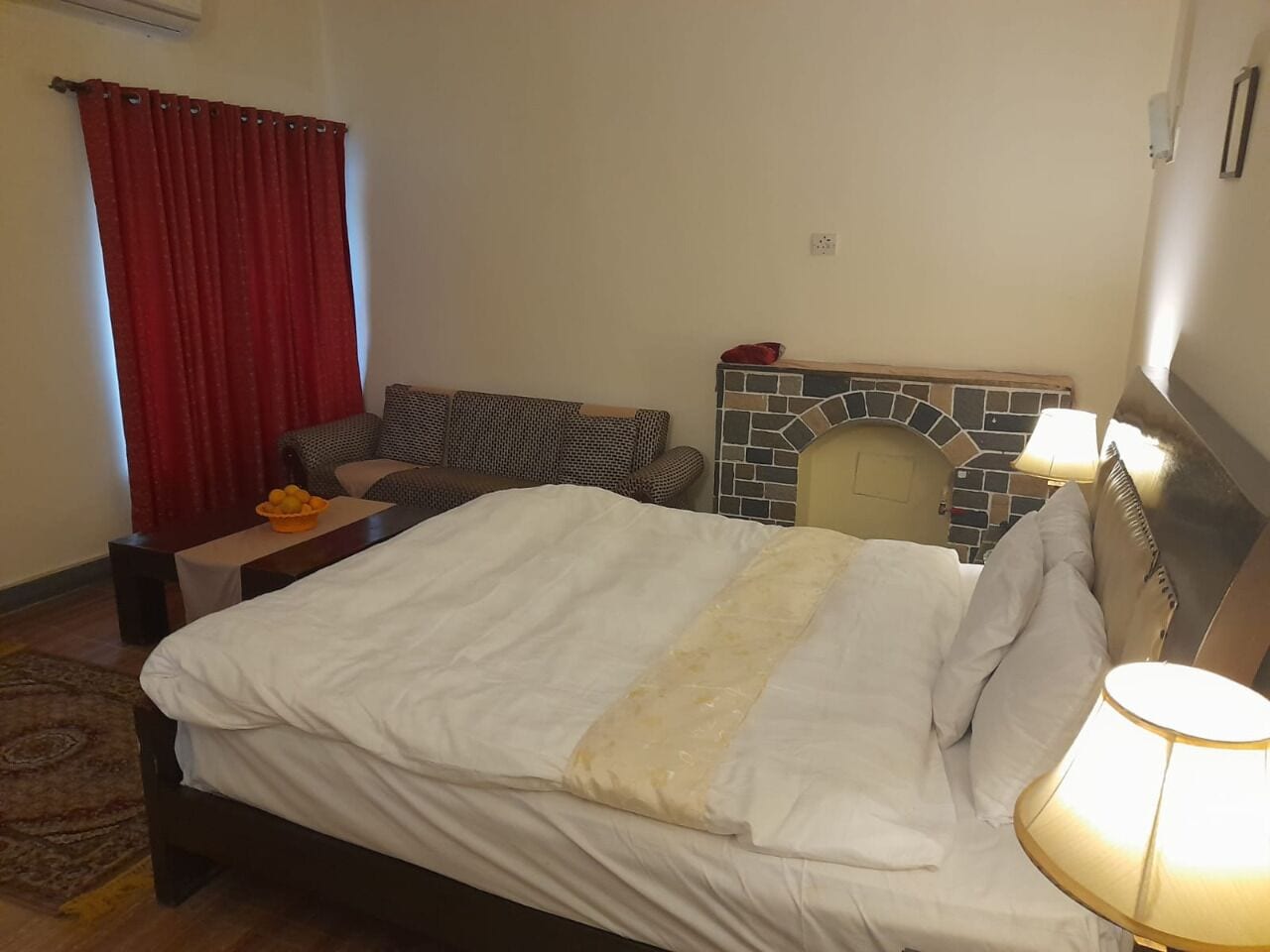 Un dormitorio con una cama, una mesita de noche, una cortina roja, un sofá estampado, una chimenea y una lámpara.