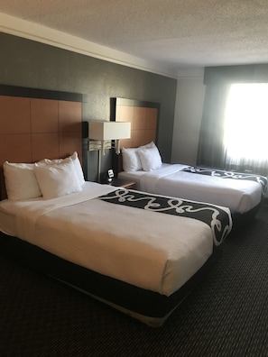 Une chambre d’hôtel avec deux lits, une tête de lit, une table de chevet avec une lampe et une fenêtre avec des rideaux.
