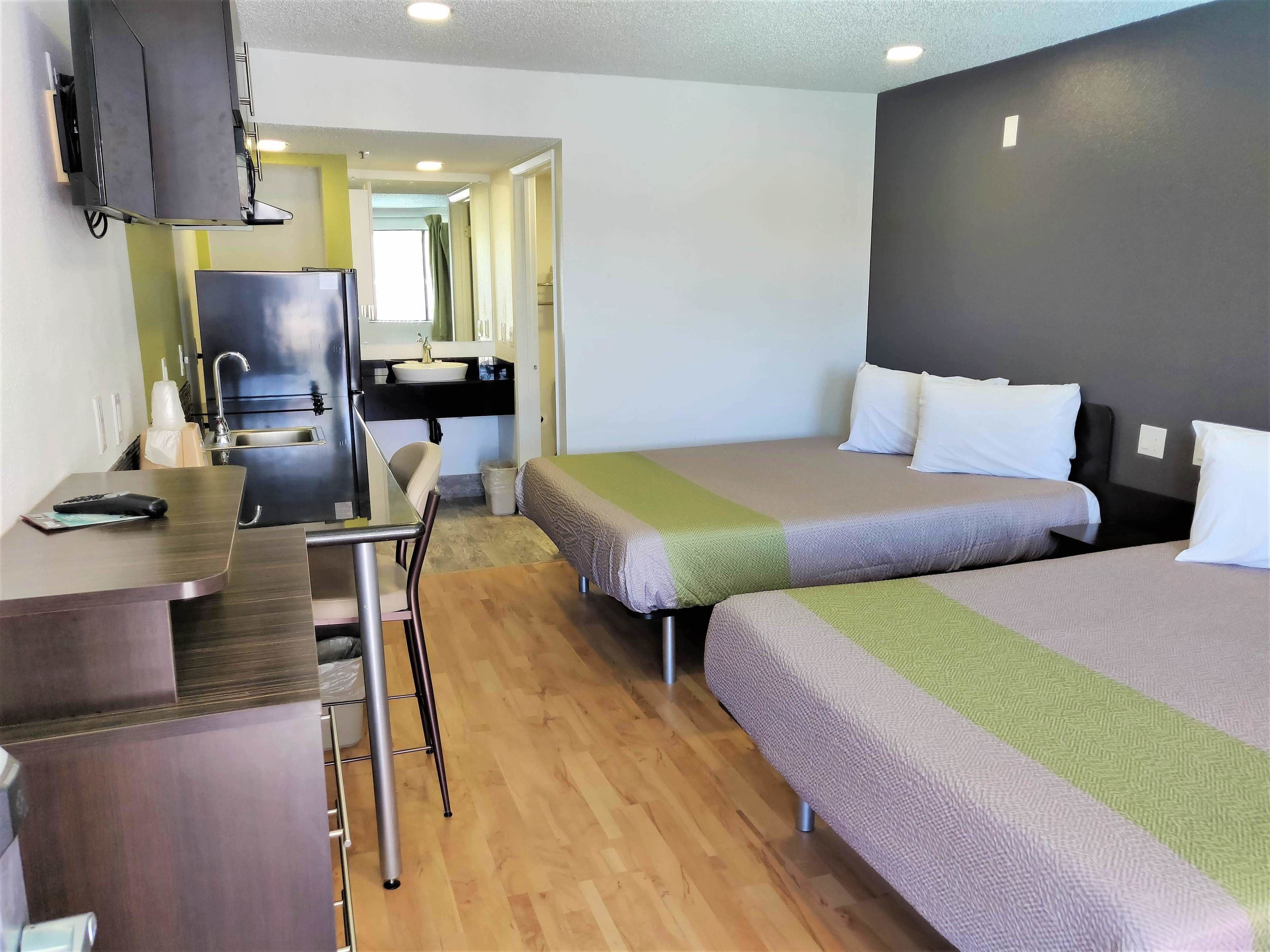 A compact hotel room with a kitchenette, two single beds, a desk with a chair, and a TV.
