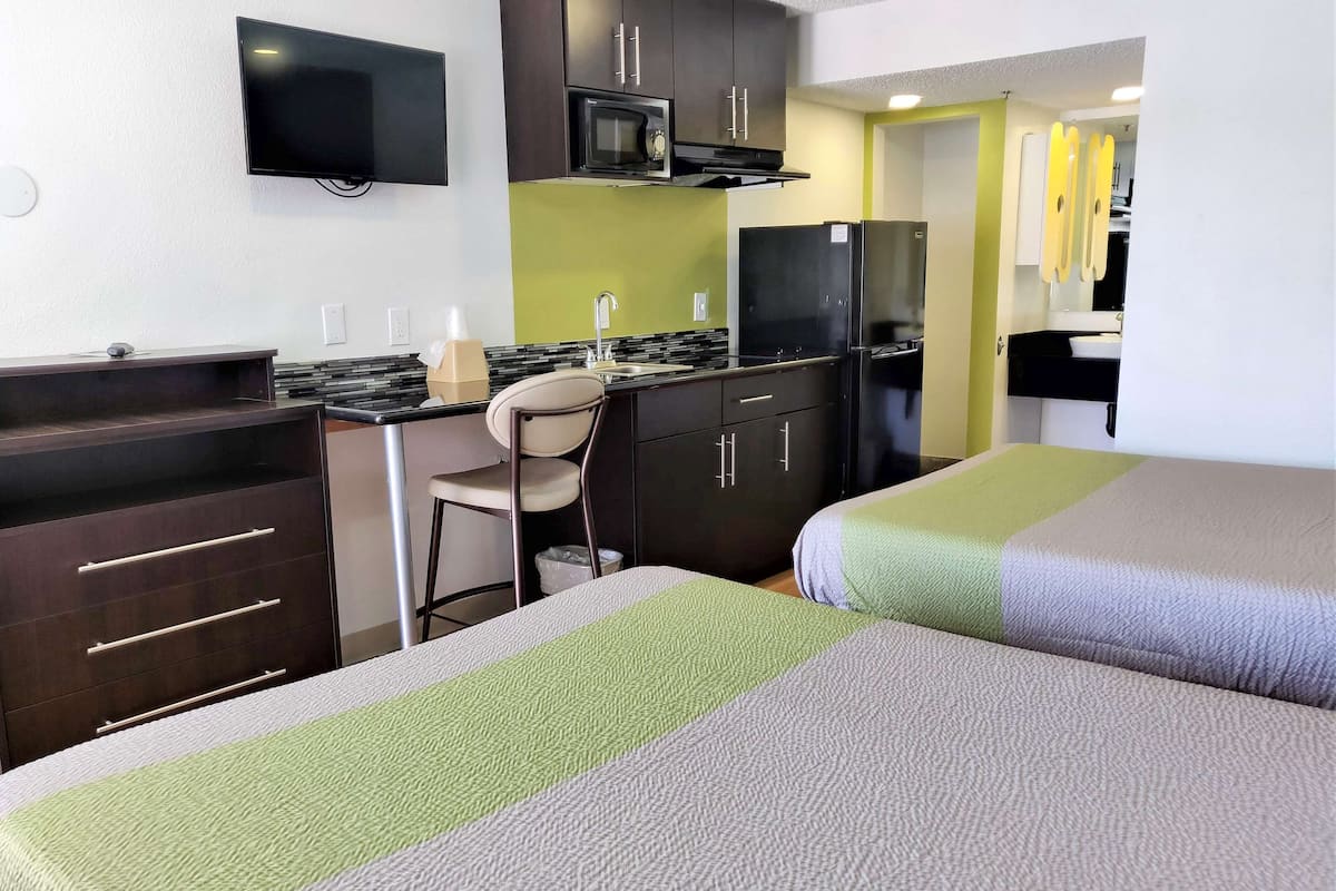 A hotel room with a kitchenette, a TV mounted on the wall, a bed with green and gray bedding, and a chair.
