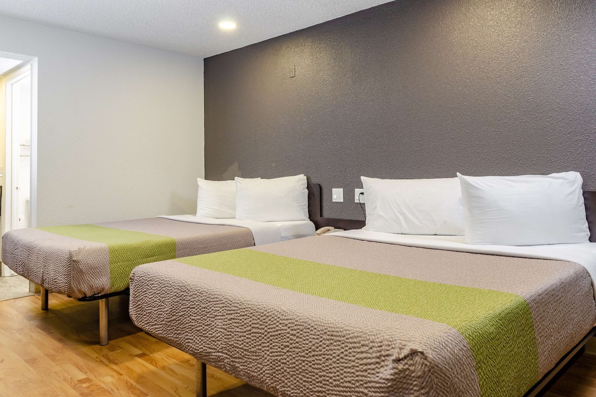A hotel room with two single beds, each with a green and beige blanket, white pillows, and a wooden floor.