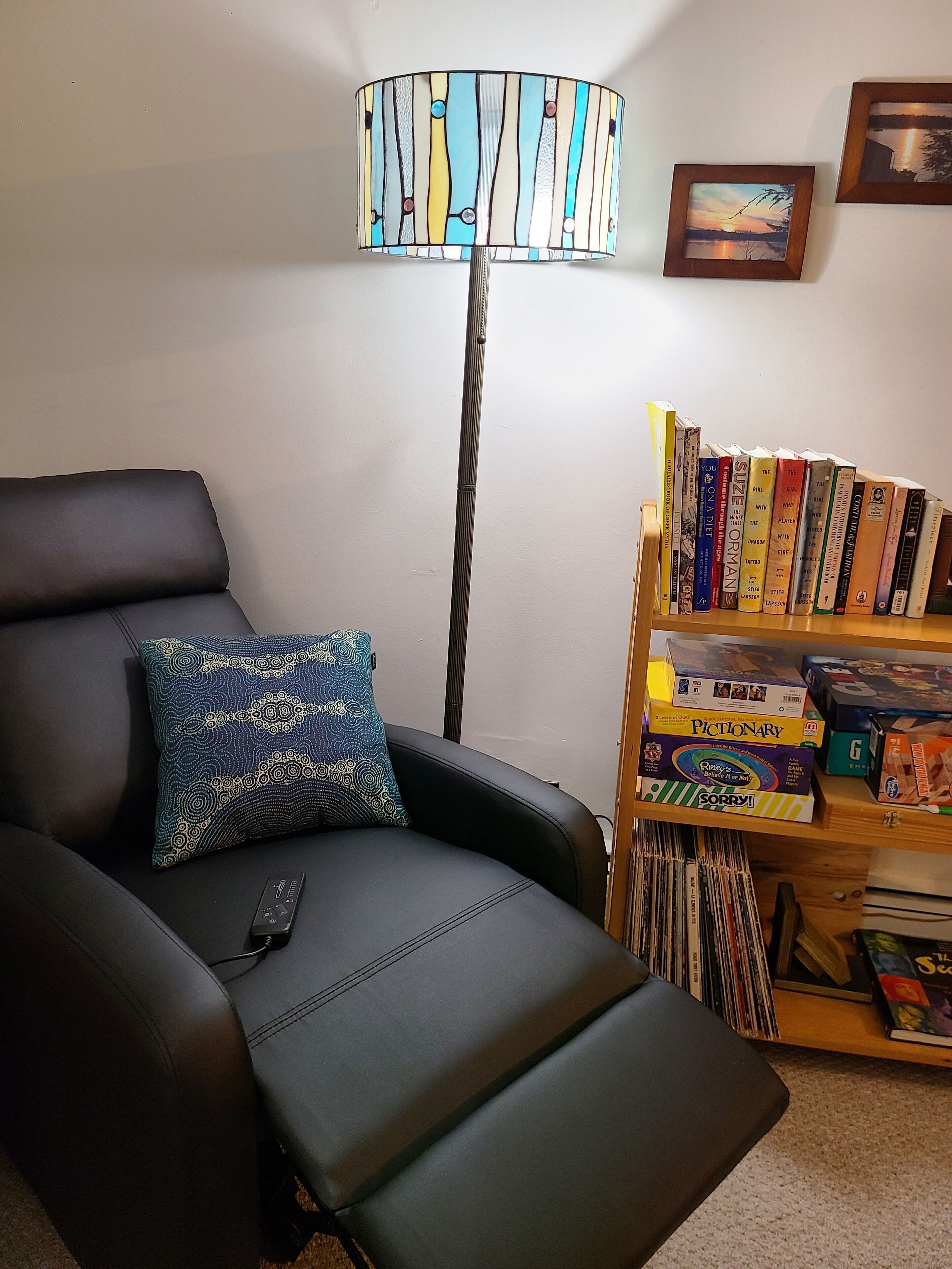 Una sala de estar con un sofá de cuero negro, un cojín estampado, una lámpara de pie, una estantería con libros y un control remoto sobre el sofá.