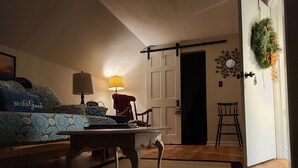 Tunnelmallinen huone, jossa on sänky, pöytä, lamppu ja tuoli. Huoneesta on ovi toiseen huoneeseen, ja seinällä on koristeellinen seppele.