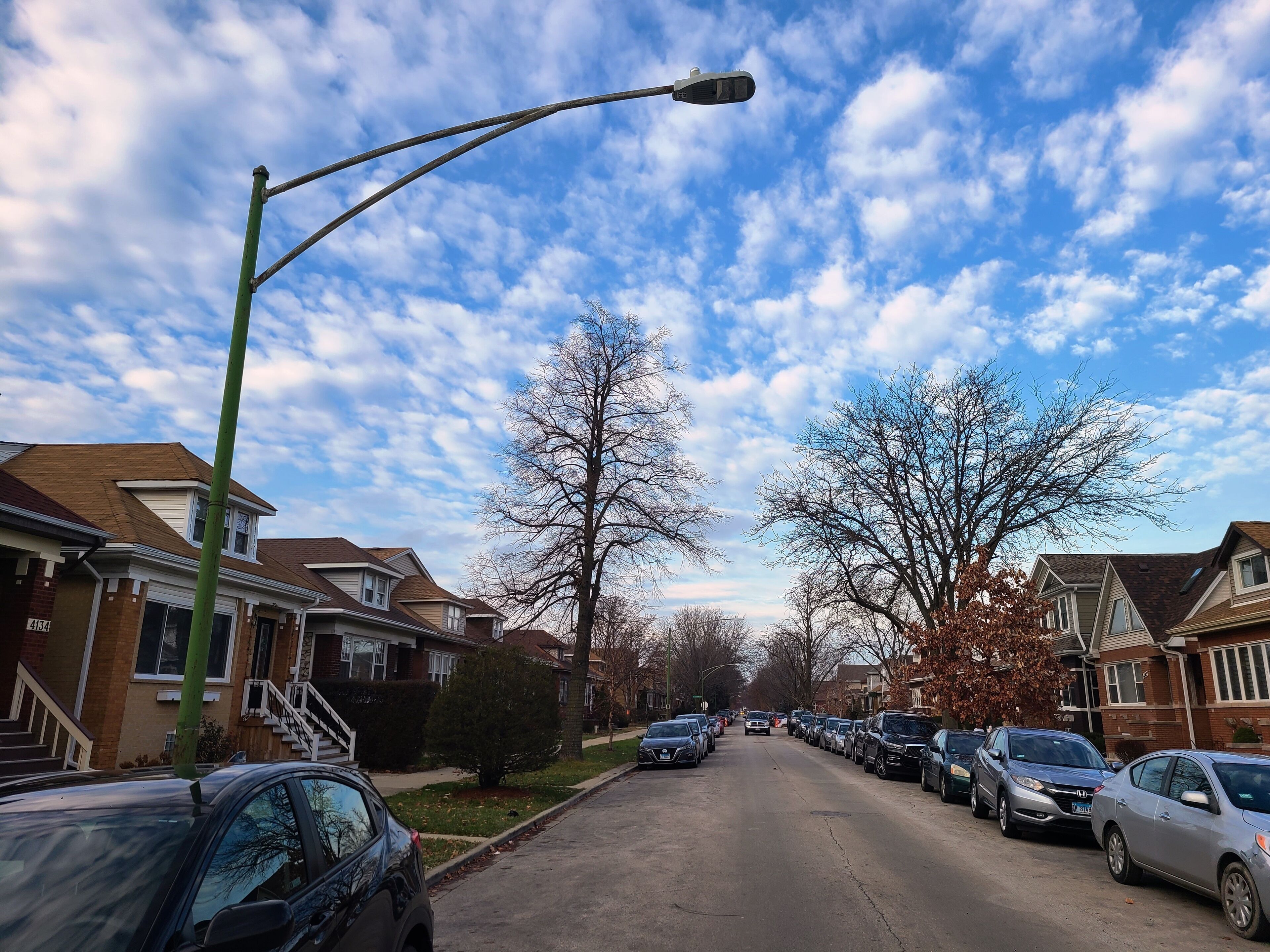 A residential street with parked cars, houses, and a clear sky.