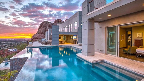 Ein modernes Hotel mit Pool, einer großen Felsformation und Blick auf die Stadt im Abendrot.