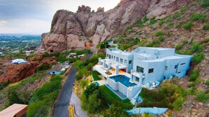 Une maison moderne à plusieurs étages, dotée d’une piscine, située sur une colline et offrant une vue sur une vallée et le paysage urbain lointain.