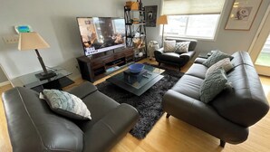 A modern living room with a flat-screen TV, leather sofas, a coffee table, and a black rug. -  N TacomaPt Ruston 2bd. 2bath retreat w/ EV charger, A/C & relaxing water views (Tacoma)