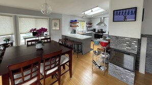 A modern kitchen with a central island, a dining area with wooden chairs, and a TV mounted on the wall. -  N TacomaPt Ruston 2bd. 2bath retreat w/ EV charger, A/C & relaxing water views (Tacoma)