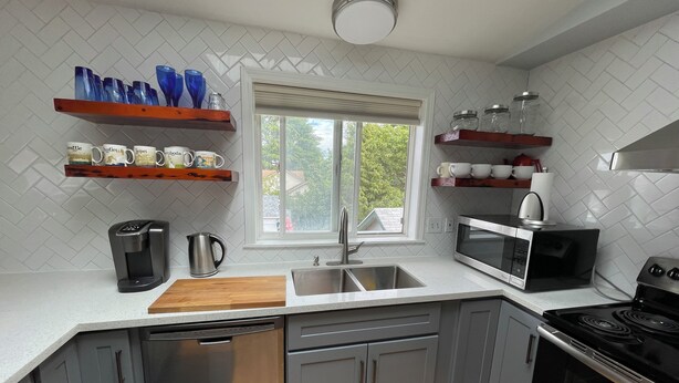 A modern kitchen with grey cabinets, a dishwasher, a stove, a microwave, and a coffee maker. There are shelves with mugs and a window with a view of greenery. -  N TacomaPt Ruston 2bd. 2bath retreat w/ EV charger, A/C & relaxing water views (Tacoma)