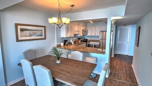 Un comedor con una mesa de madera y sillas azul claro, una cocina con armarios blancos, y un pasillo con una puerta.