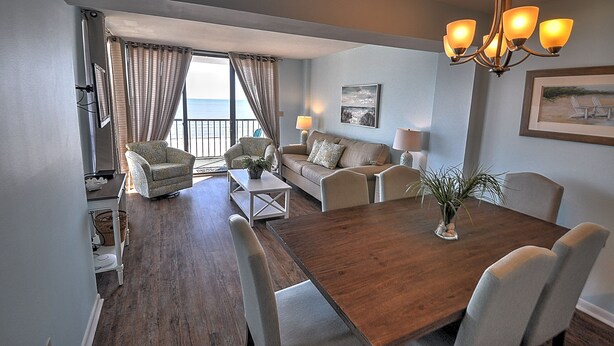 Una sala de estar con una mesa de comedor de madera, sofás color beige y vista al mar a través de amplios ventanales.