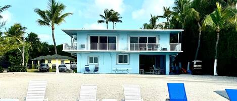 A two-story beach house with a balcony, multiple windows, and a covered patio area.