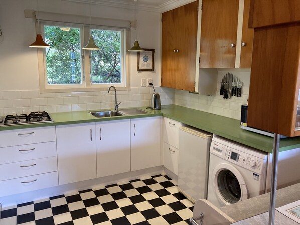 A kitchen with white cabinets, a green countertop, a black and white checkered floor, a window with a view of trees, and a Bosch washing machine. - Comfortable retro bach, close to everything (Anzac Bay)